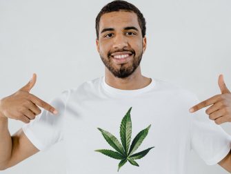 man with short black hair and beard wearing and pointing at his white tshirt with a cannabis leaf design
