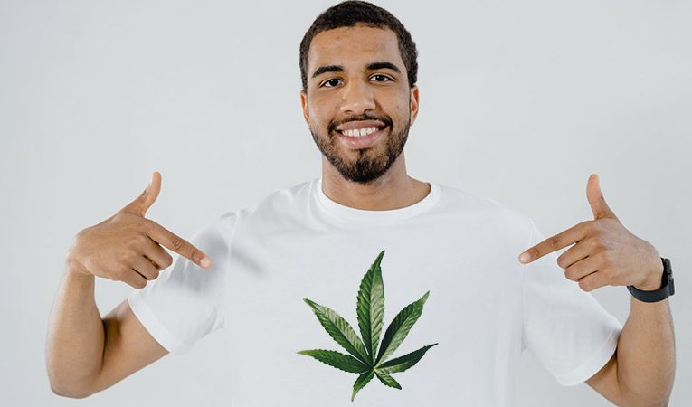 man with short black hair and beard wearing and pointing at his white tshirt with a cannabis leaf design